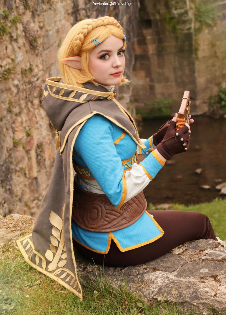 A person dressed in a blue and brown costume, resembling a character from a fantasy game, with long blonde hair and pointed ears, sitting on a rock by a riverbank and holding a small object in their hand.