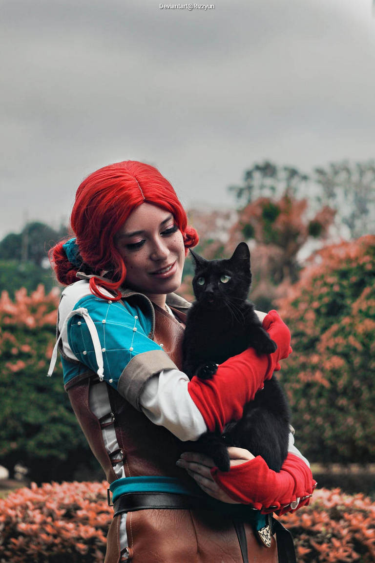 A person with red hair wearing a colorful costume and red gloves holds a black cat in a park, surrounded by autumn foliage.