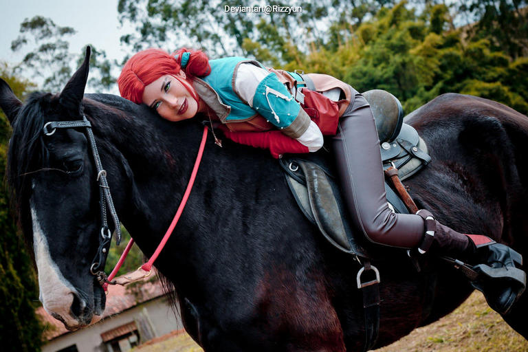 A person with red hair in a colorful outfit is leaning affectionately on a black horse while sitting in a saddle in a natural setting.
