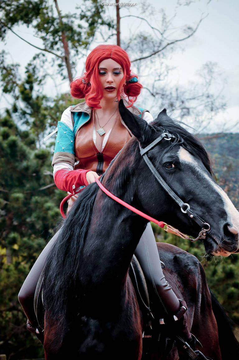 A person with red hair styled in curls is riding a black horse. They are wearing a costume with a brown top and blue accents. The background features trees and greenery.