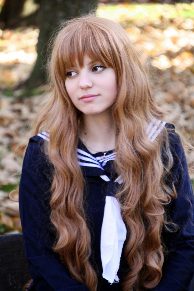 A young woman with long, wavy brown hair wearing a navy blue school uniform with a white collar. She is sitting outdoors, looking thoughtfully to the side, with fallen leaves in the background.
