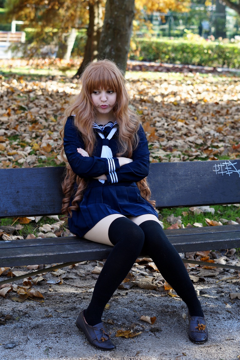 A girl with long brown hair sitting on a bench in a park, wearing a school uniform with a sailor collar and knee-high socks, surrounded by autumn leaves.