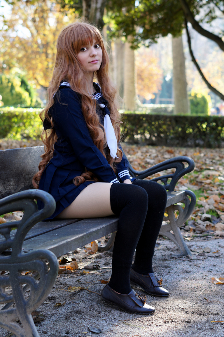 A young woman with long, wavy brown hair wearing a school uniform with a navy blue jacket and a white necktie, seated on a park bench surrounded by autumn foliage.