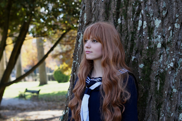 A young woman with long, wavy, reddish-brown hair stands against a large tree, wearing a navy blue outfit with a white collar and ribbon. She gazes thoughtfully into the distance with a serene expression, surrounded by a natural outdoor setting during autumn.