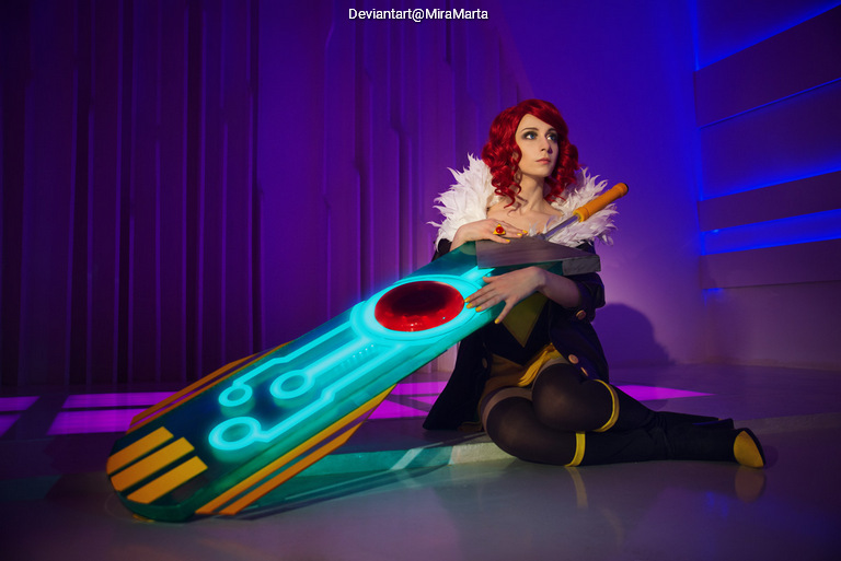 A person in a colorful costume poses with a large, illuminated weapon against a purple backdrop. They have curly red hair and an elaborate outfit featuring feathers.
