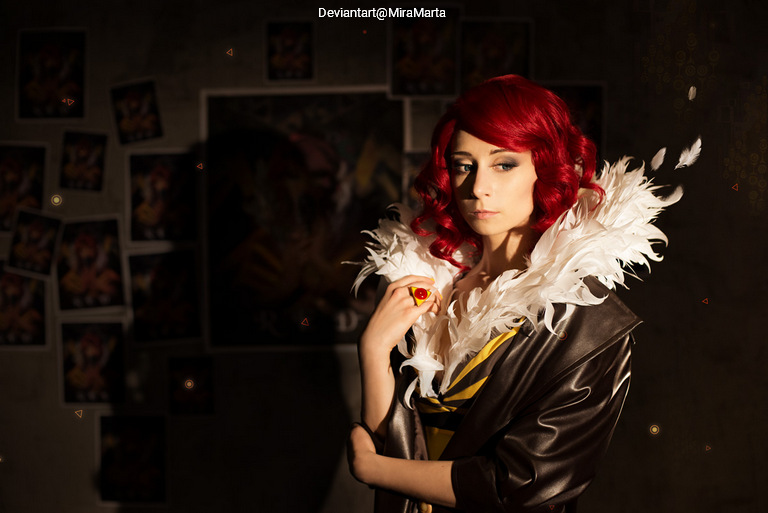 A woman with red curly hair wearing a feathered collar and a dark jacket, posing thoughtfully in a dimly lit room decorated with posters.