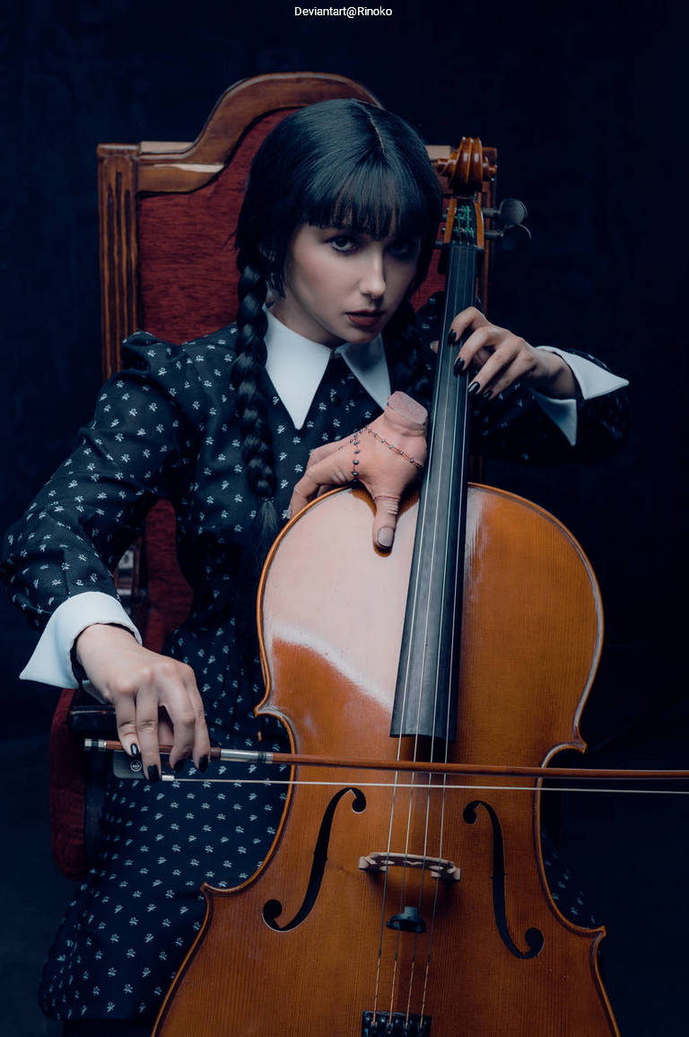 A girl with black braided hair in a polka dot dress playing a cello, sitting on a wooden chair against a dark background.