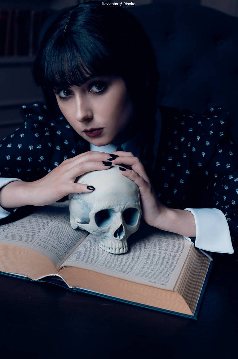 A young woman with dark hair and pale skin sits at a table, resting her hands on a human skull placed on an open book.