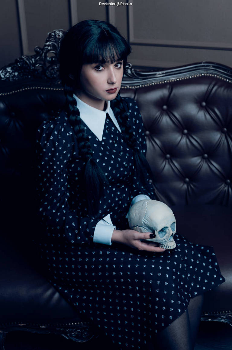 A young woman with dark hair styled in braids, wearing a polka dot dress with a white collar, sitting on a vintage brown sofa while holding a human skull.