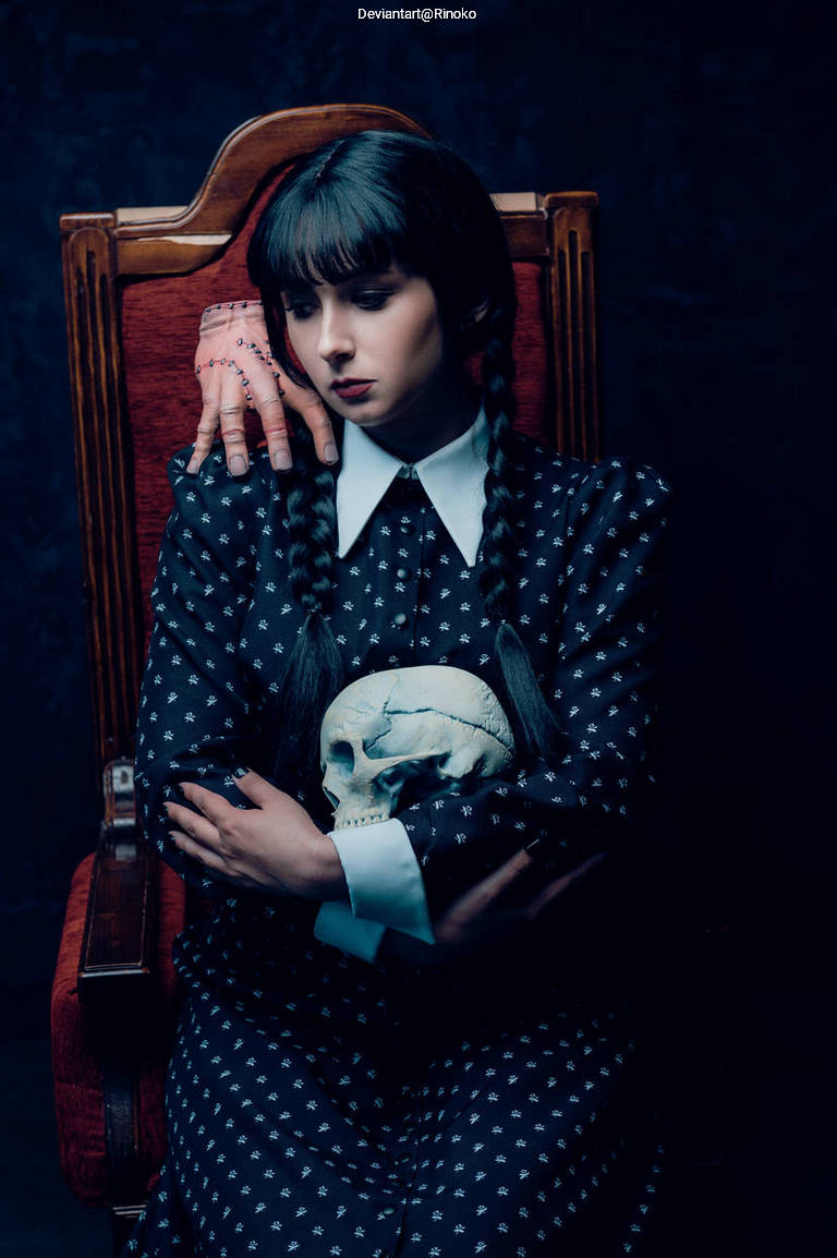 A young woman with dark hair styled in braids sits in an ornate chair, holding a skull in her lap. She wears a gothic dress with white patterns and has a somber expression. A hand is seen resting on the back of her chair, adding an eerie element to the scene.