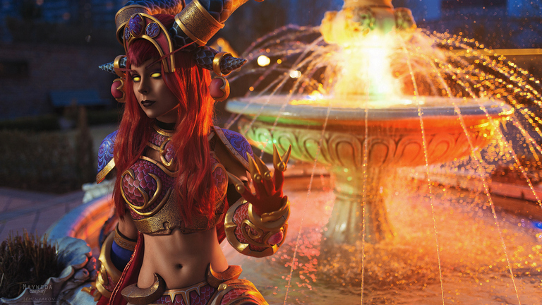 A woman in elaborate fantasy costume with red hair and gold accents poses near a colorful fountain at dusk.