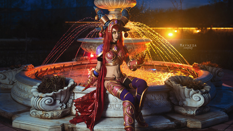 A cosplayer dressed in elaborate armor and accessories sits near a fountain, with colorful lights illuminating the scene at dusk.