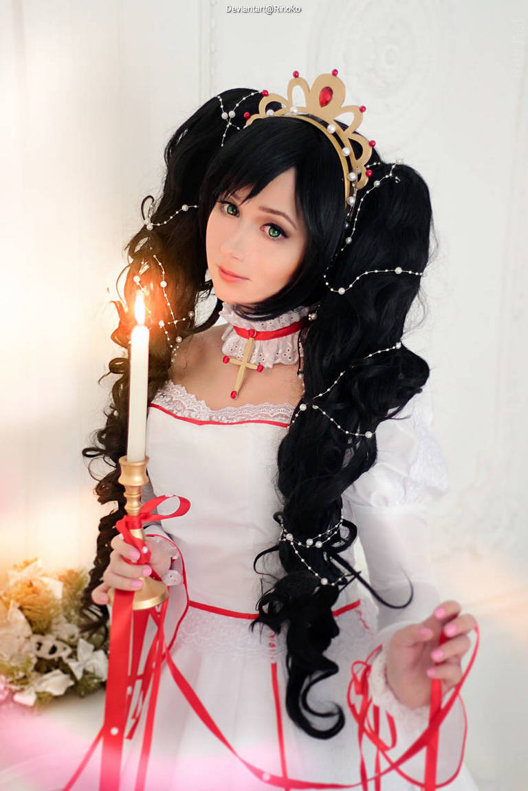 A person in a dramatic white dress with red accents, holding a candle and adorned with a crown, long dark hair decorated with beads and ribbons.