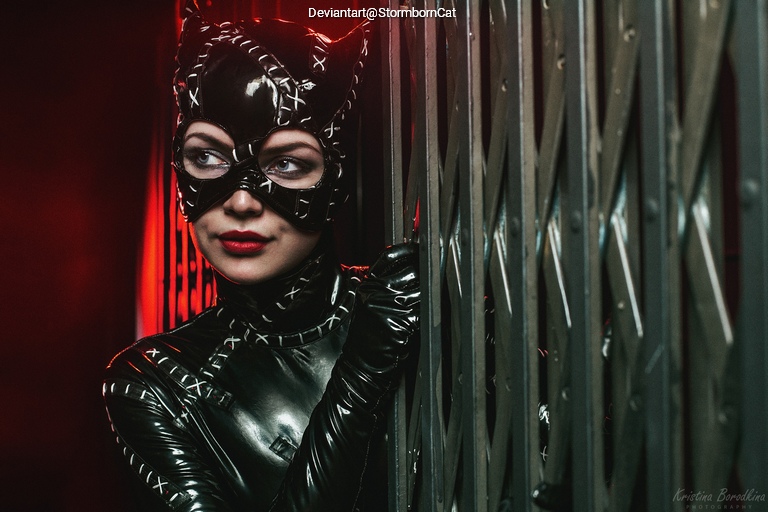 A woman dressed as a cat character, wearing a black latex costume and mask, poses next to an elevator door with a dramatic red background.