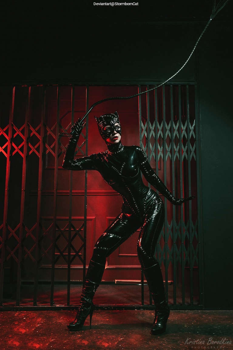 A person dressed in a shiny black catsuit with cat-like ears, holding a whip, posed dramatically in front of a dark background with red lighting.