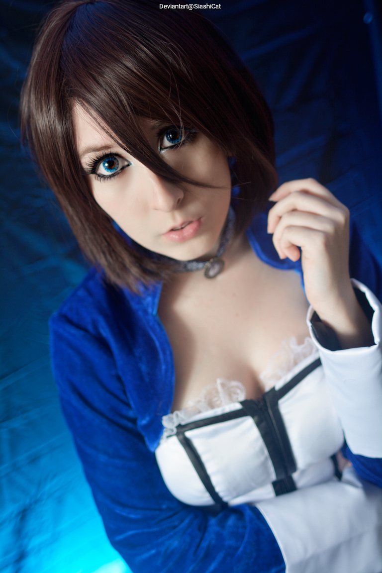 Close-up portrait of a person with short brown hair and blue eyes wearing a blue costume with a white top, set against a blue background.