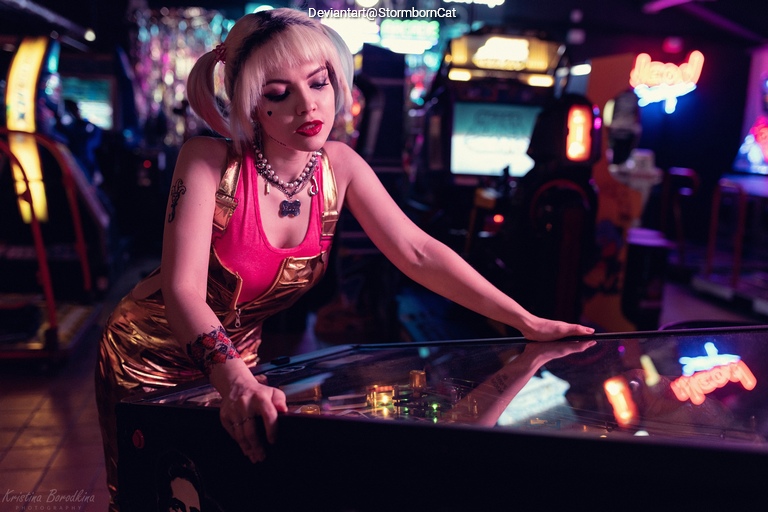 A young woman with blonde hair styled in pigtails, wearing a pink top and metallic gold overalls, leans over a pinball machine in a vibrant arcade, surrounded by colorful neon lights.