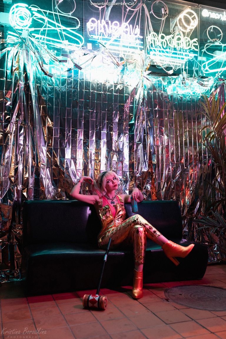 A person in shiny golden attire sits on a black couch in a colorful, neon-lit environment, surrounded by reflective decor, holding a drink and a toy mallet.