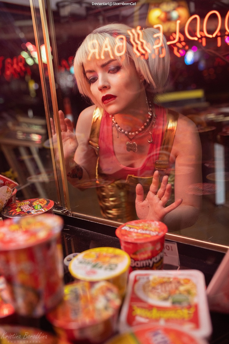 A young woman with short blonde hair gazes longingly at a display of instant noodles through a glass counter, with neon signs reflected in the glass.