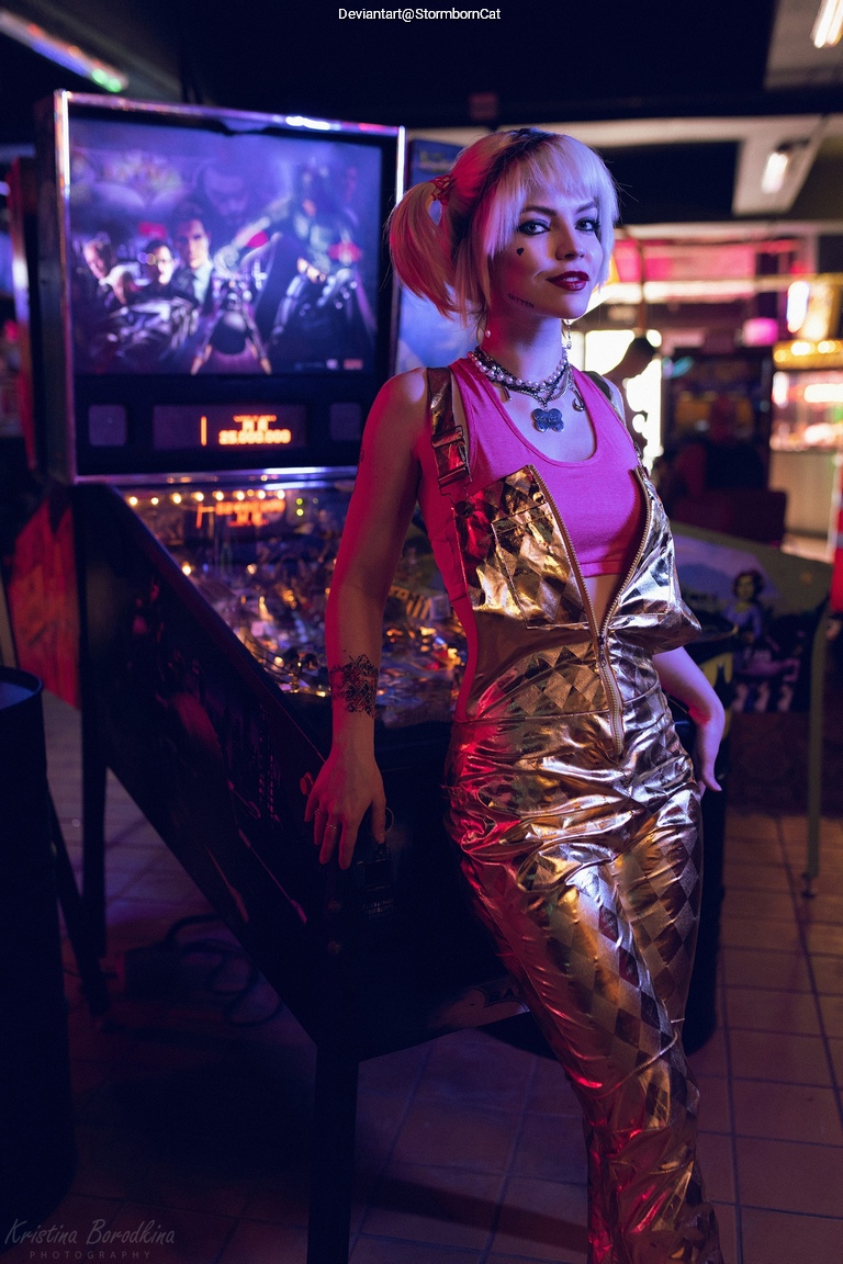 A person dressed in a shiny gold outfit poses near a pinball machine in a dimly lit arcade.