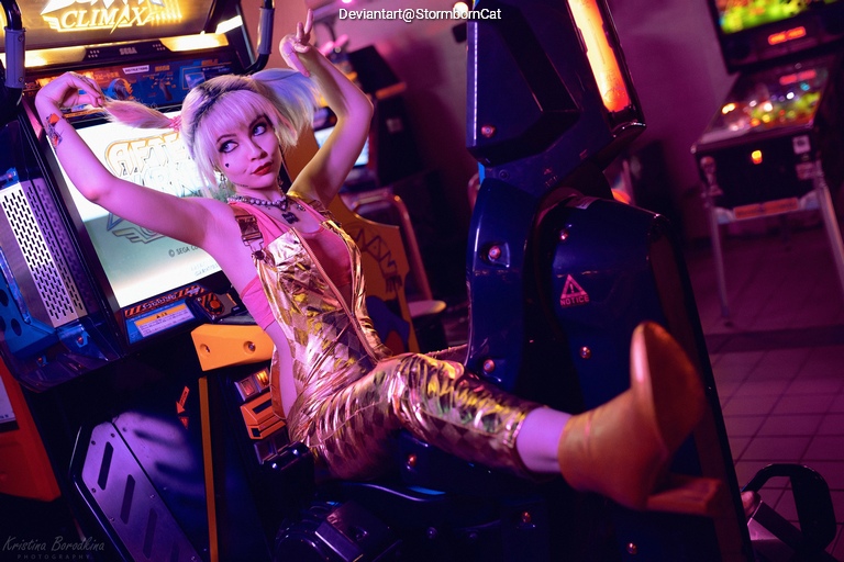 A person wearing a shiny gold jumpsuit and high heels poses playfully in an arcade, with colorful arcade machines in the background.
