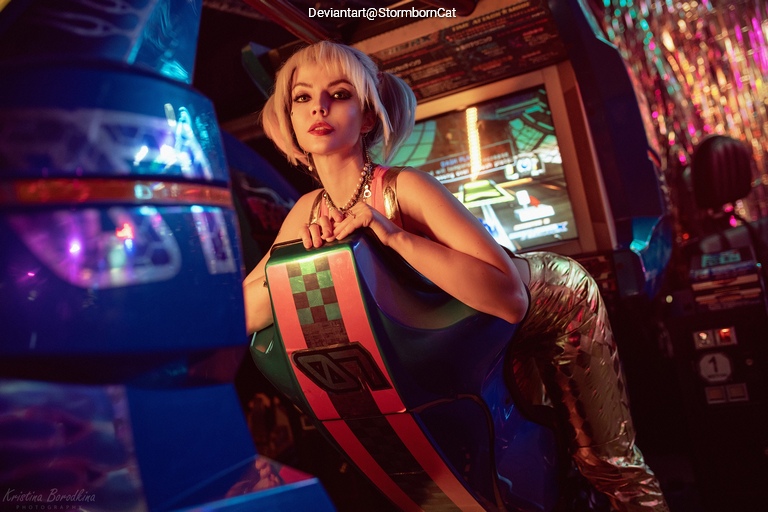 A person with blonde hair styled in pigtails poses in front of a colorful arcade game, wearing a shiny outfit and jewelry.
