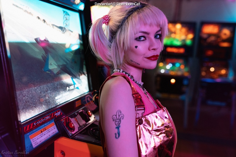 A person with pink hair and bold makeup poses near an arcade game, showcasing a playful and edgy style.