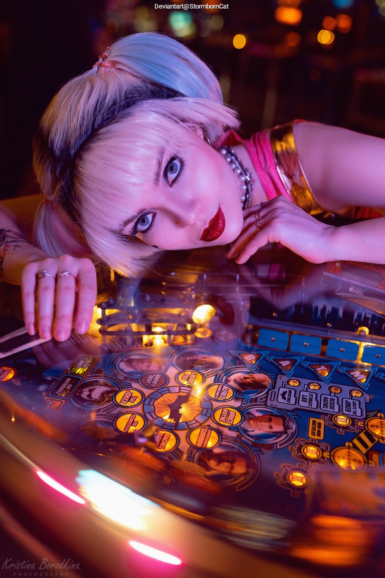 A close-up of a person with pastel-colored hair leaning over a pinball machine, showcasing vibrant lights and game graphics.
