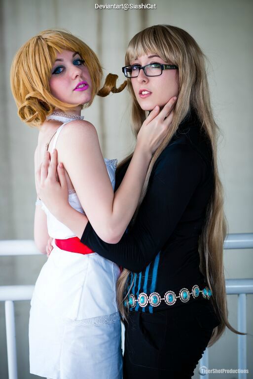 Two individuals in cosplay embracing each other, one with blonde hair in curls wearing a white dress with a red belt, and the other with long brown hair wearing a black outfit with a decorative belt.