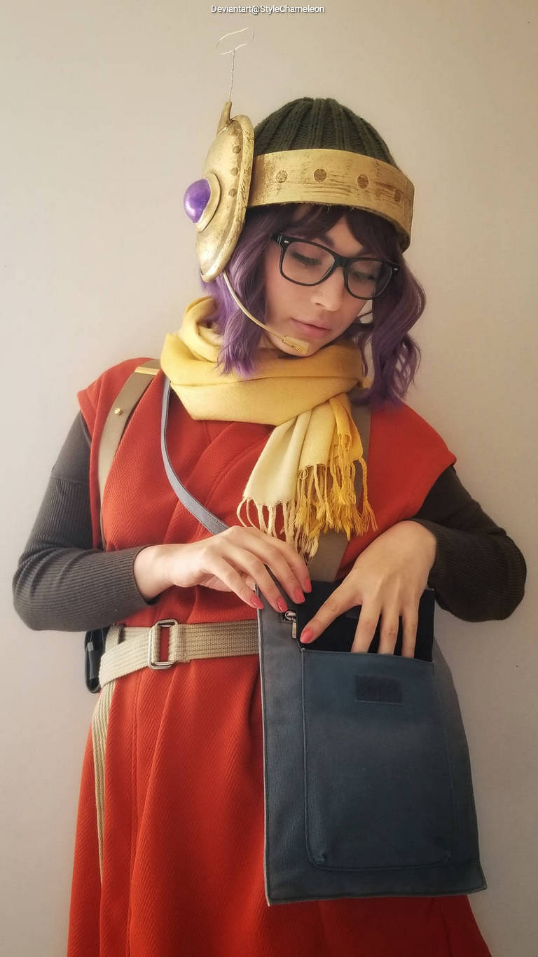 A person wearing a colorful costume with a helmet featuring a large decorative element and a purple gem, along with glasses and a yellow scarf, is reaching into a blue purse.