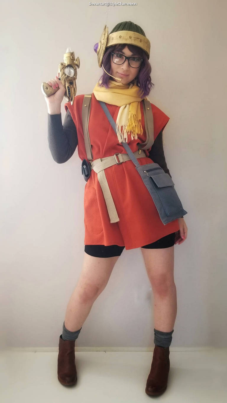 A person dressed in a colorful character costume, holding a prop gun and striking a pose. They have purple hair, glasses, and are wearing a bright orange top with a scarf and a satchel.