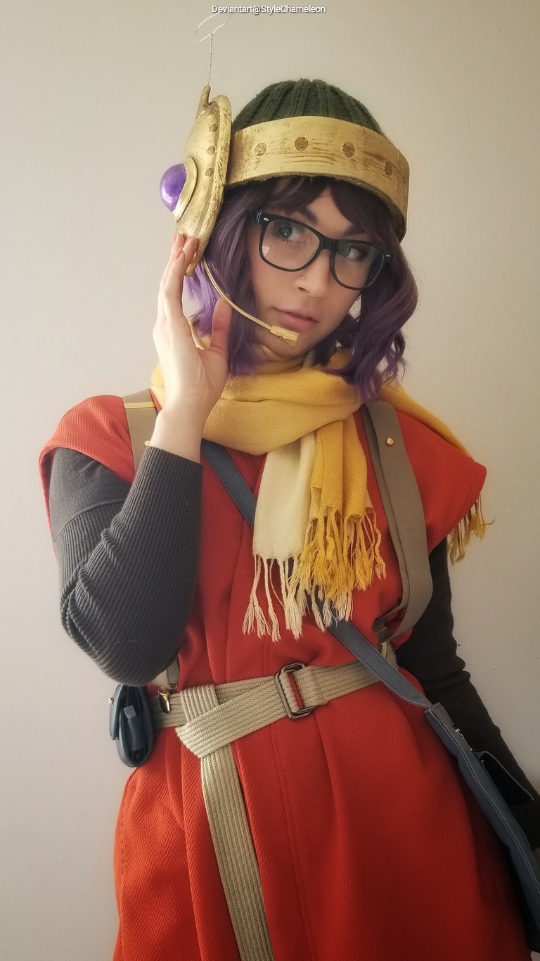 A person wearing a colorful costume and headpiece, holding a headset to their ear, with glasses and a playful expression against a neutral background.