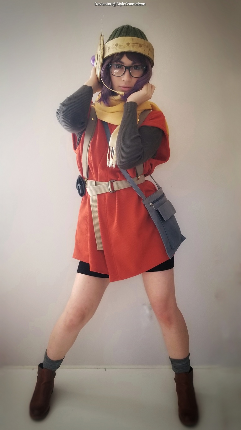 A person dressed in a colorful costume, featuring an orange top, beige belt, and a green hat, striking a pose against a light background.