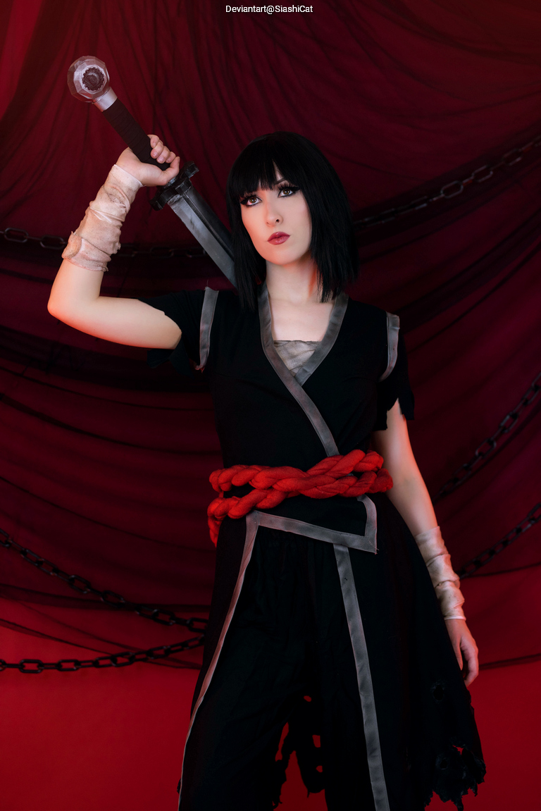 A person with short black hair, dressed in a dark outfit with red accents, poses confidently while holding a sword above their shoulder against a dramatic red and black backdrop.
