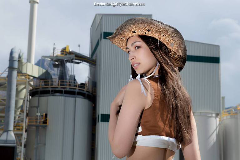 A person wearing a stylish cowboy hat and a brown outfit, standing in front of industrial structures.