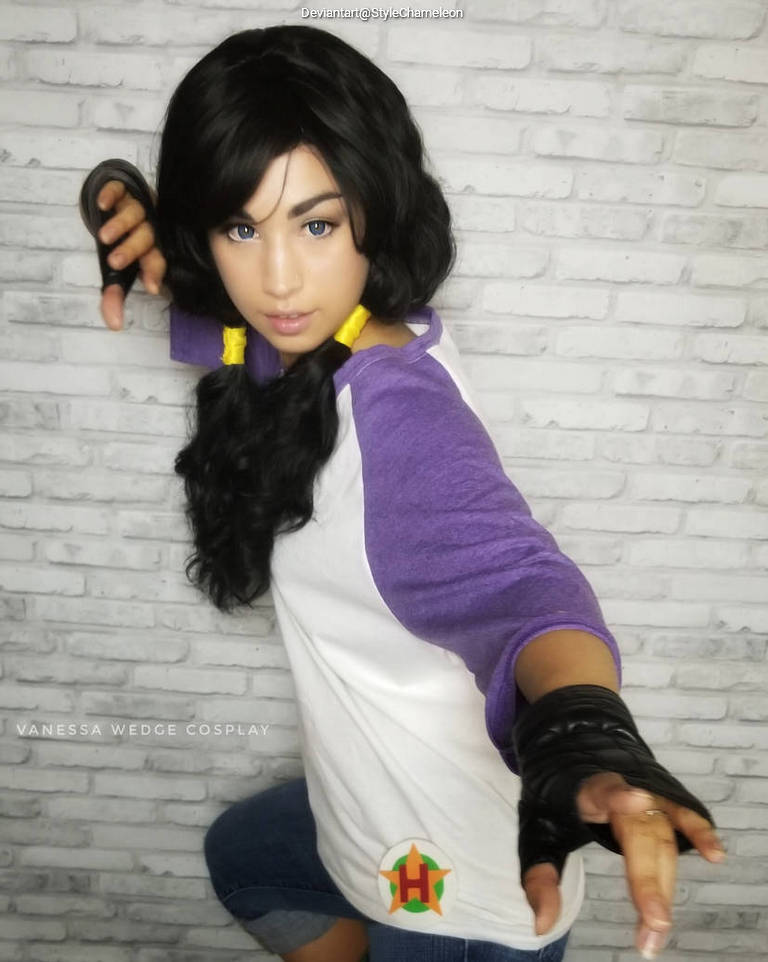 A person cosplaying with a black wig and purple shirt, striking a dynamic pose against a brick wall background.