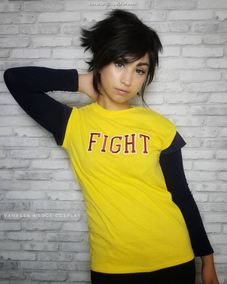 A person posing with one hand on their hip, wearing a yellow t-shirt with the word 'FIGHT' in bold red letters and a black long-sleeve shirt underneath. The background features a white brick wall.