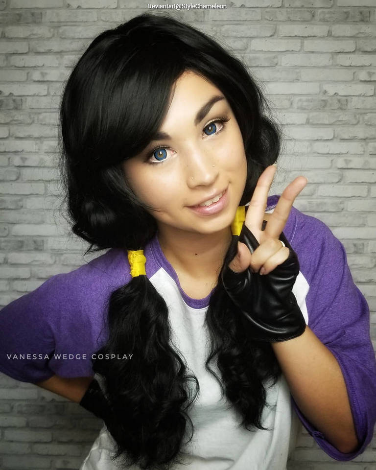 A person with long black hair styled in pigtails, wearing a purple and white shirt, and black gloves, smiling and making a peace sign with their hand against a brick wall background.