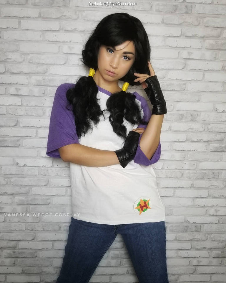 A person in a white and purple oversized shirt poses against a brick wall, wearing black gloves and styled wavy hair with yellow accessories.