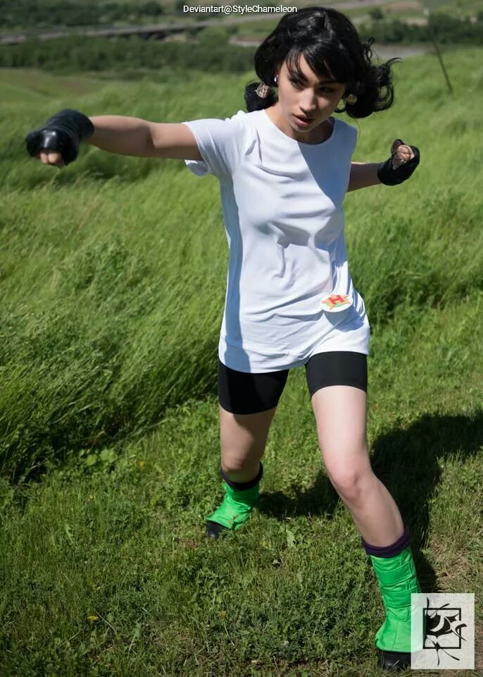 A person in a white shirt and black shorts poses dynamically in a grassy field, showcasing a martial arts stance with fists raised, wearing green boots and gloves.