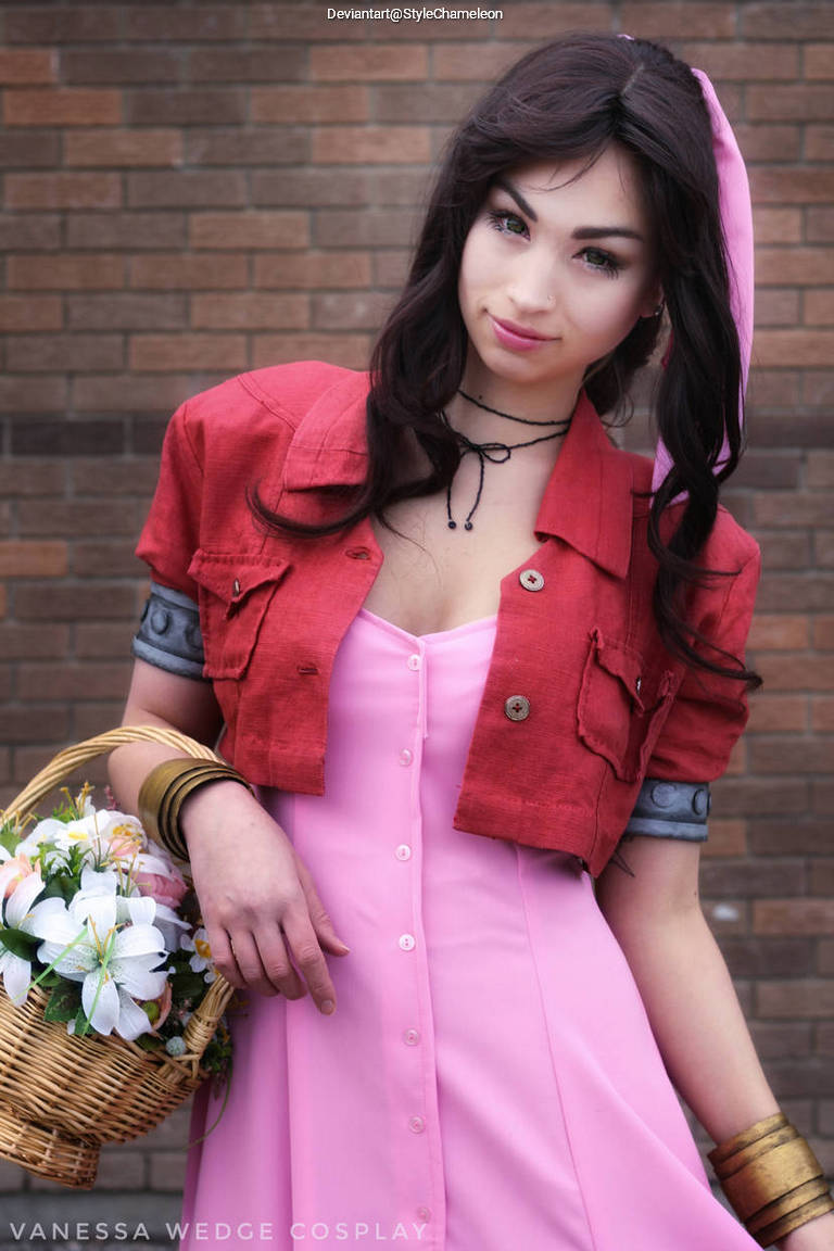 A woman dressed in a pink dress and a red cropped jacket, holding a basket of flowers. She has long dark hair and subtle accessories, standing against a brick wall.