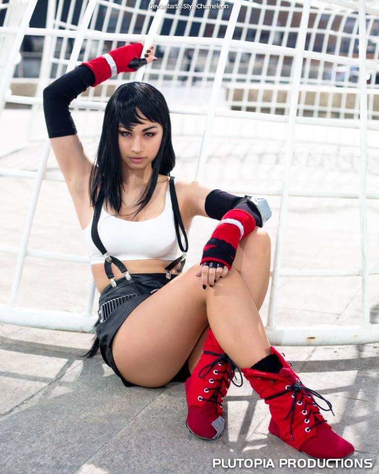 A young woman posing in a stylish outfit with a white top, black shorts, and red knee-high boots. She has long dark hair and is sitting against a white structure, striking a confident pose.