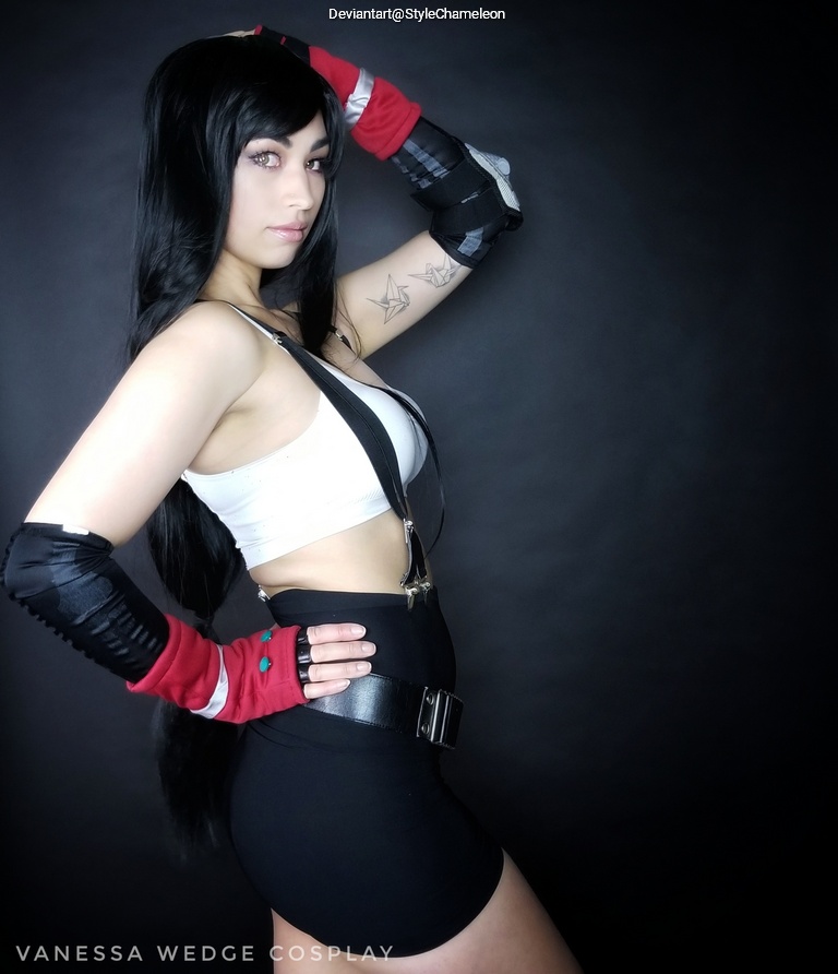 A person in cosplay, wearing a cropped top and a fitted black skirt, with long black hair and gloves. The background is dark, highlighting the costume details.