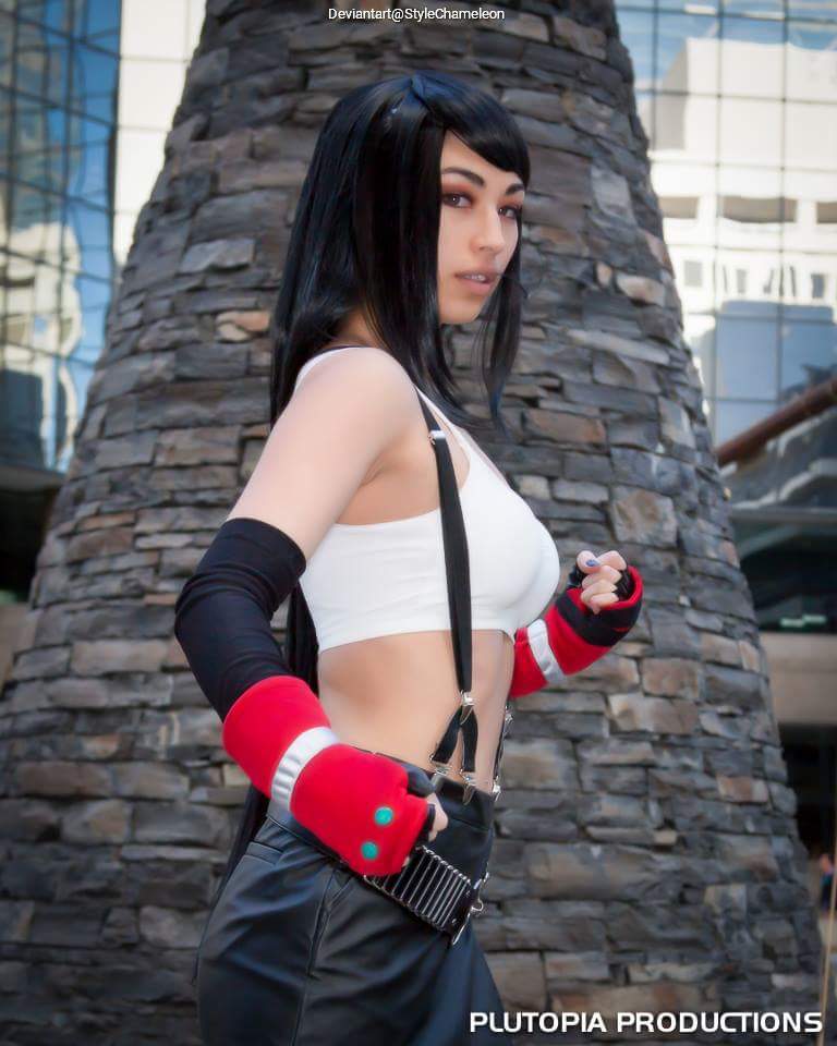 Person in a cosplay outfit with a black wig, wearing a white top and black pants, standing in front of a stone backdrop with a modern building in the background.