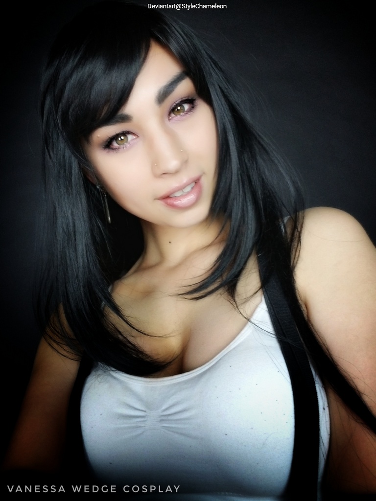 Close-up portrait of a person with long black hair, wearing a white tank top and black suspenders, smiling at the camera against a dark background.
