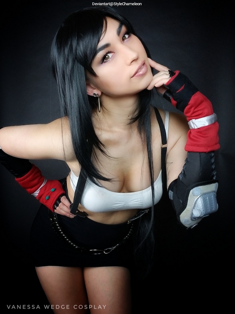 A person posing in a costume, featuring a fitted white top, a short black skirt, and gloves with intricate designs. They have long black hair and are smiling, with one hand resting on their chin.