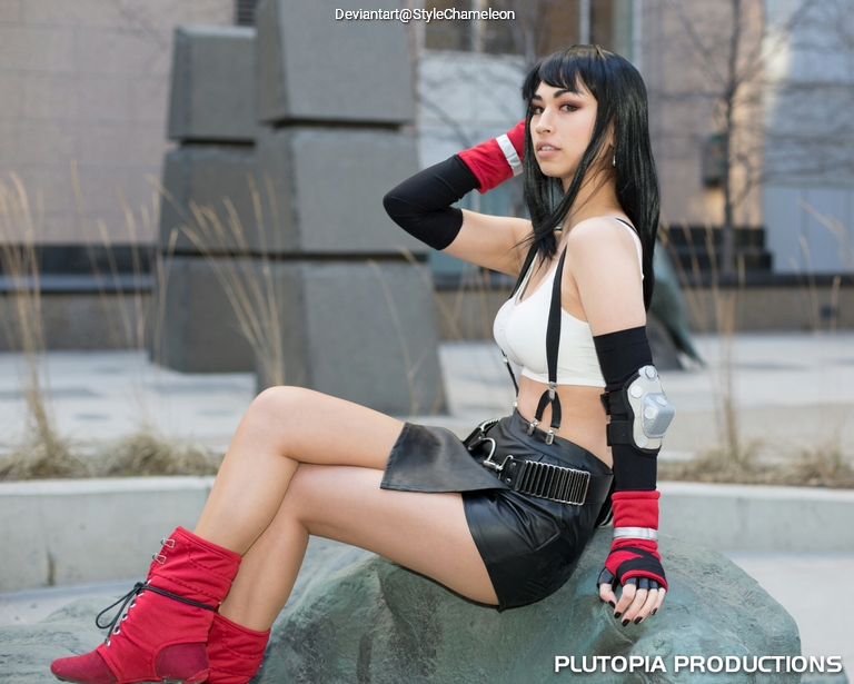 A female cosplayer in a black skirt and white crop top, sitting on a rock with a city background. She has long black hair and is wearing red boots and gloves.