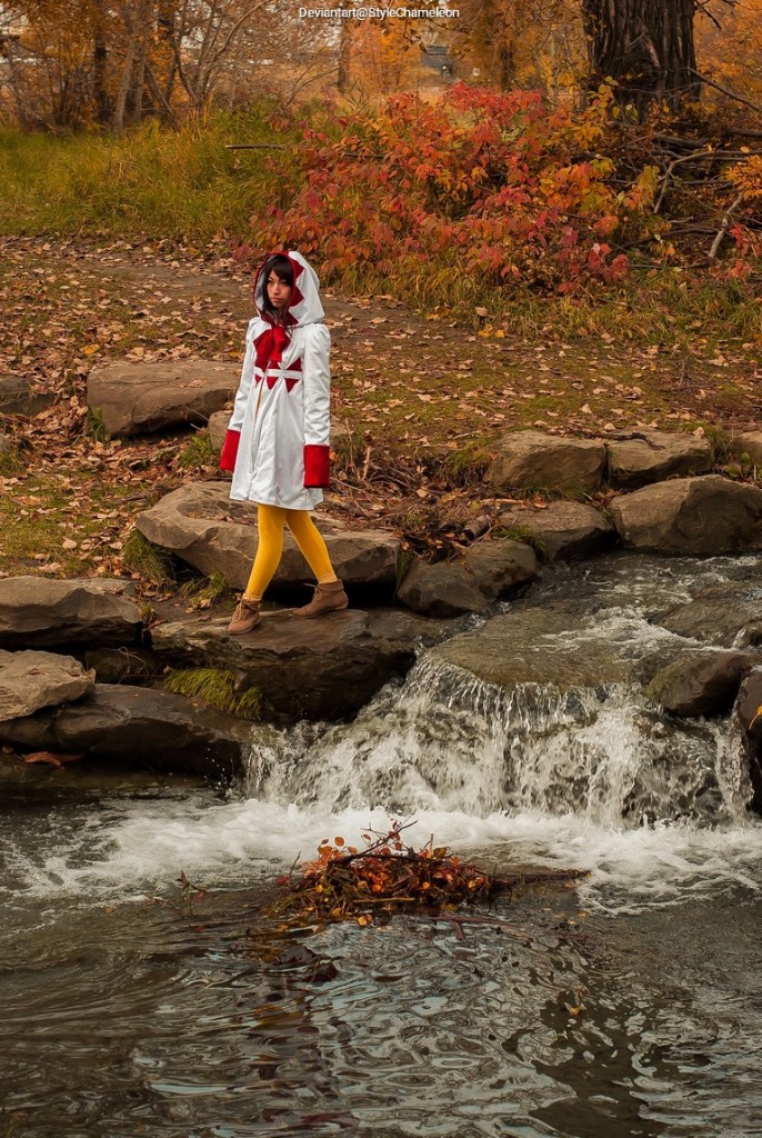A person dressed in a white coat with a red hood and yellow leggings stands on rocks beside a flowing stream, surrounded by autumn foliage.