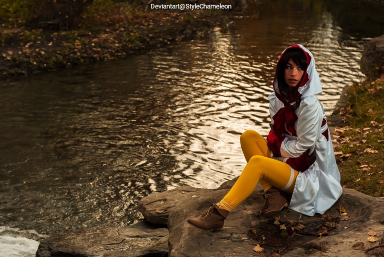 A person sitting on a rock by a stream, wearing a white and red hooded cloak with yellow leggings, surrounded by autumn foliage.