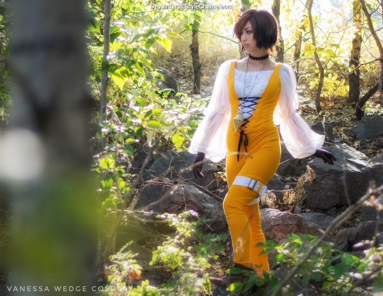 A person dressed in a yellow and white cosplay outfit stands amidst a natural setting with green foliage and rocks.